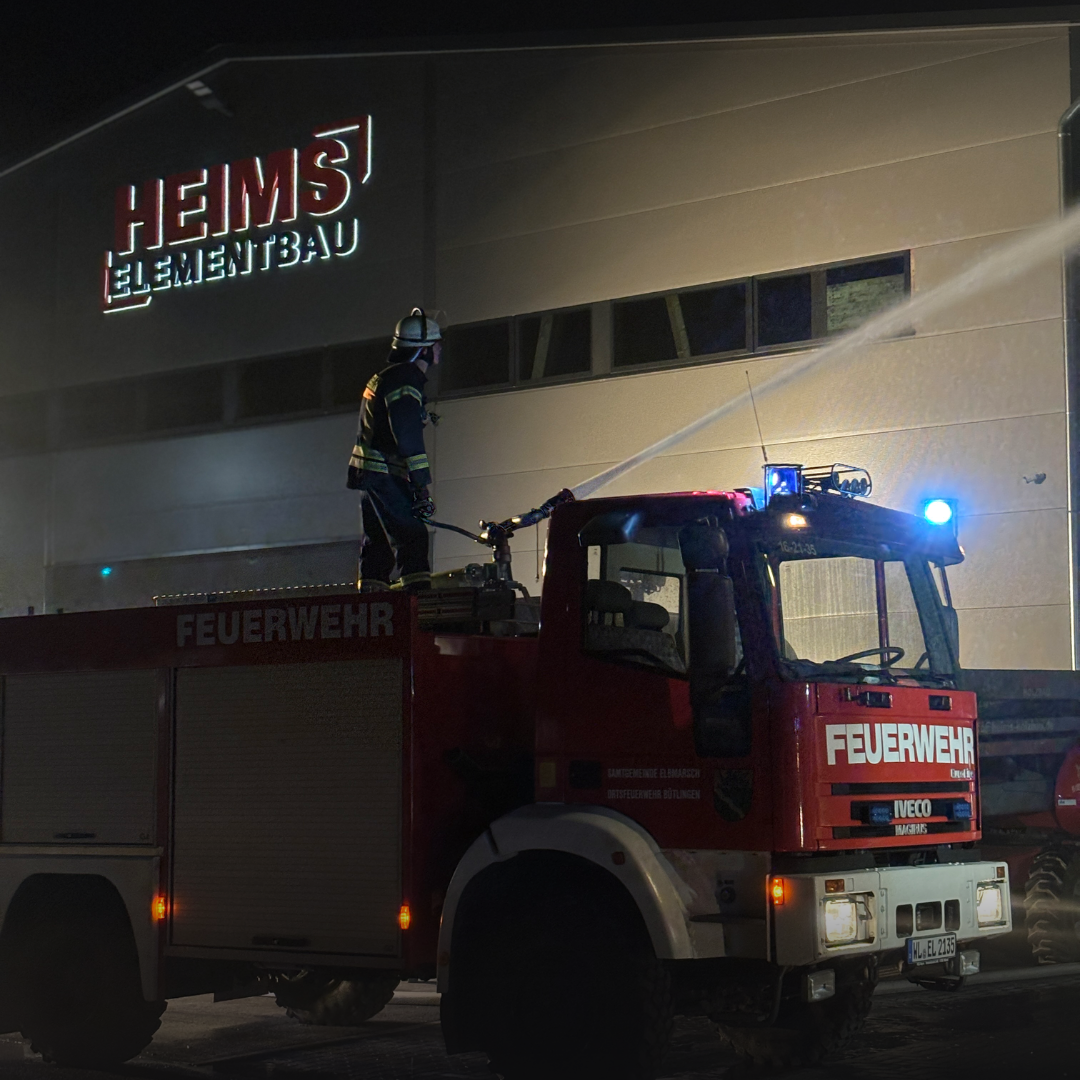 Feuerwehrmann steht nachts auf dem Dach eines Löschfahrzeugs vor einer Lagerhalle, ein Wasserstrahl trifft die Fassade.