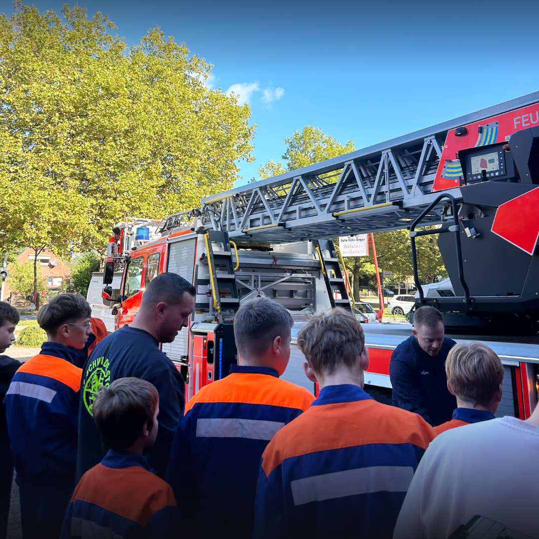 Eine Gruppe Jugendlicher in Jugendfeuerwehruniform steht draußen vor einer Drehleiter, ein Feuerwehrmann erklärt den Bedienstand.