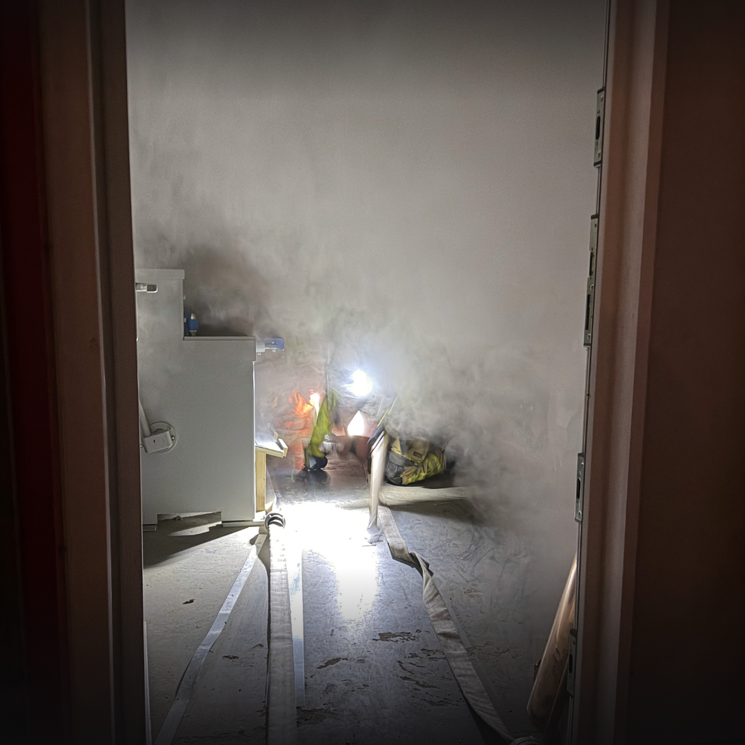 Blick durch eine Tür in einen verrauchten Raum, eine Einsatzkraft mit Lampe kniet am Boden, ein Schlauch führt in den Raum.