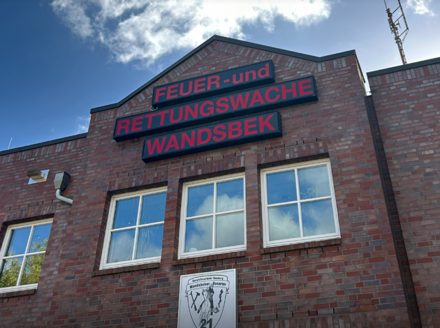 Rotes Backsteingebäude mit dem Schriftzug Feuer- und Rettungswache Wandsbek unter blauem Himmel, darunter ein Wappenschild mit der Nummer 21.