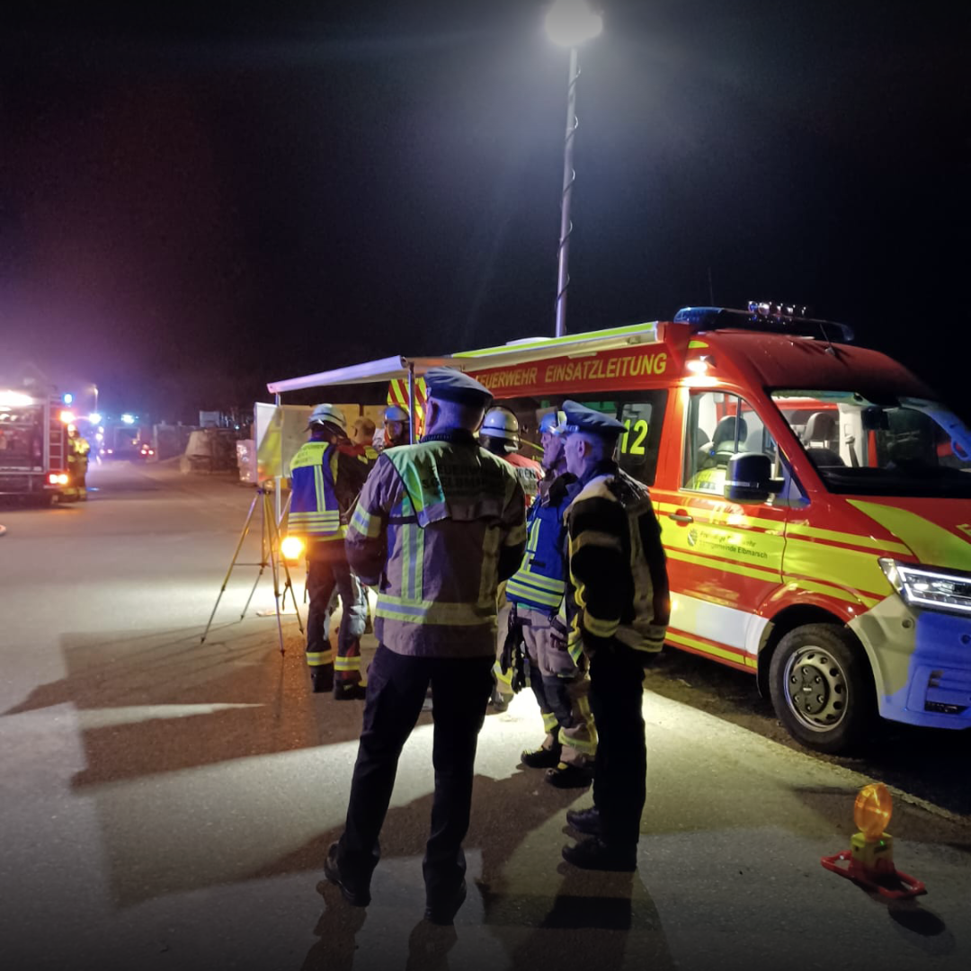 Drei Feuerwehrleute stehen nachts im Gespräch vor einem Einsatzleitwagen, im Hintergrund weitere Fahrzeuge mit Blaulicht.