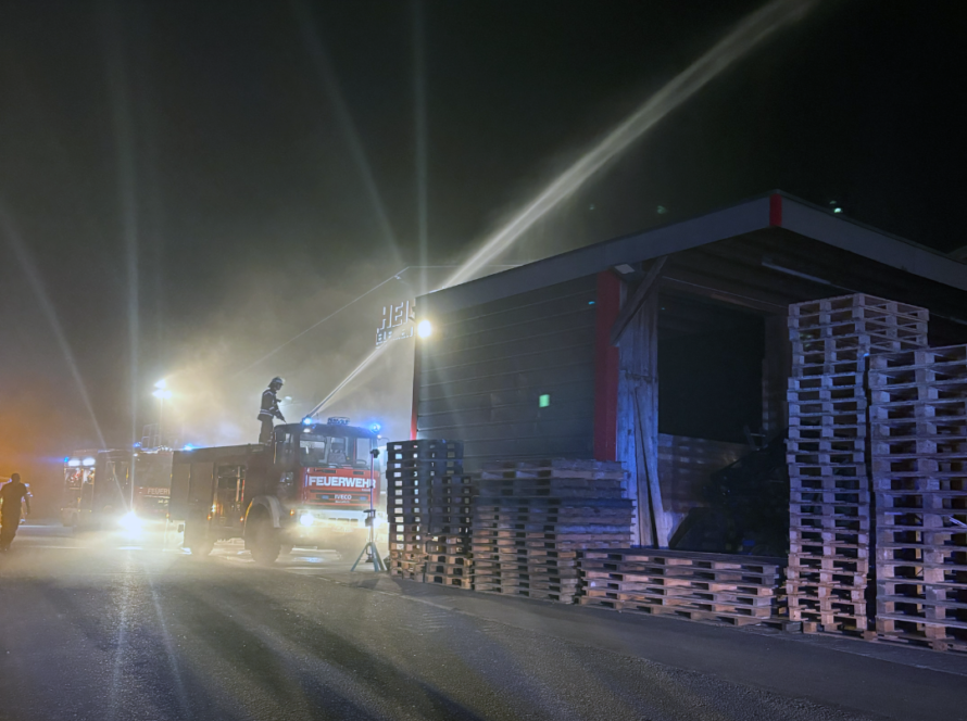 Wasserstrahl über einem überdachten Palettenlager bei Nacht, links ein Löschfahrzeug mit Einsatzkraft auf dem Dach.