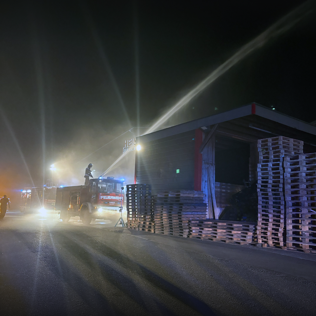 Wasserstrahl über einem überdachten Palettenlager bei Nacht, links ein Löschfahrzeug mit Einsatzkraft auf dem Dach.