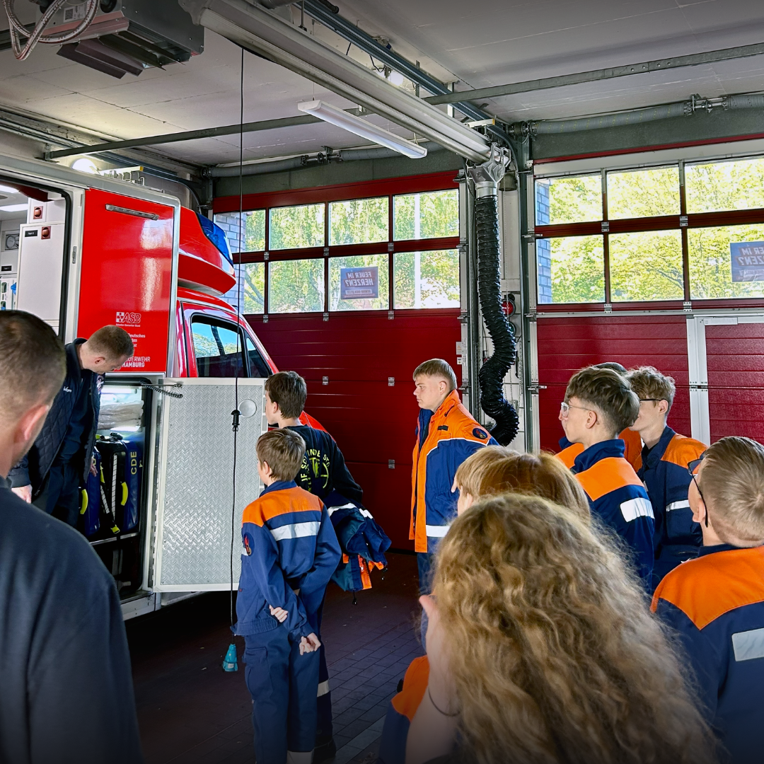 Eine Gruppe Jugendlicher steht in der Fahrzeughalle vor einem Einsatzfahrzeug mit geöffneten Geräteräumen, ein Feuerwehrmann erklärt die Ausrüstung.