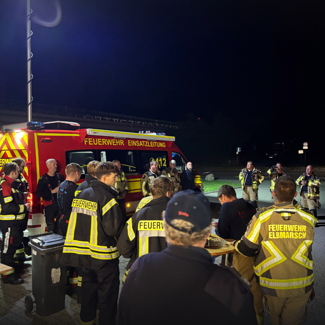 Gruppe von Feuerwehrleuten in Einsatzkleidung steht nachts vor einem Einsatzleitwagen, einige tragen gelbe Funktionswesten.