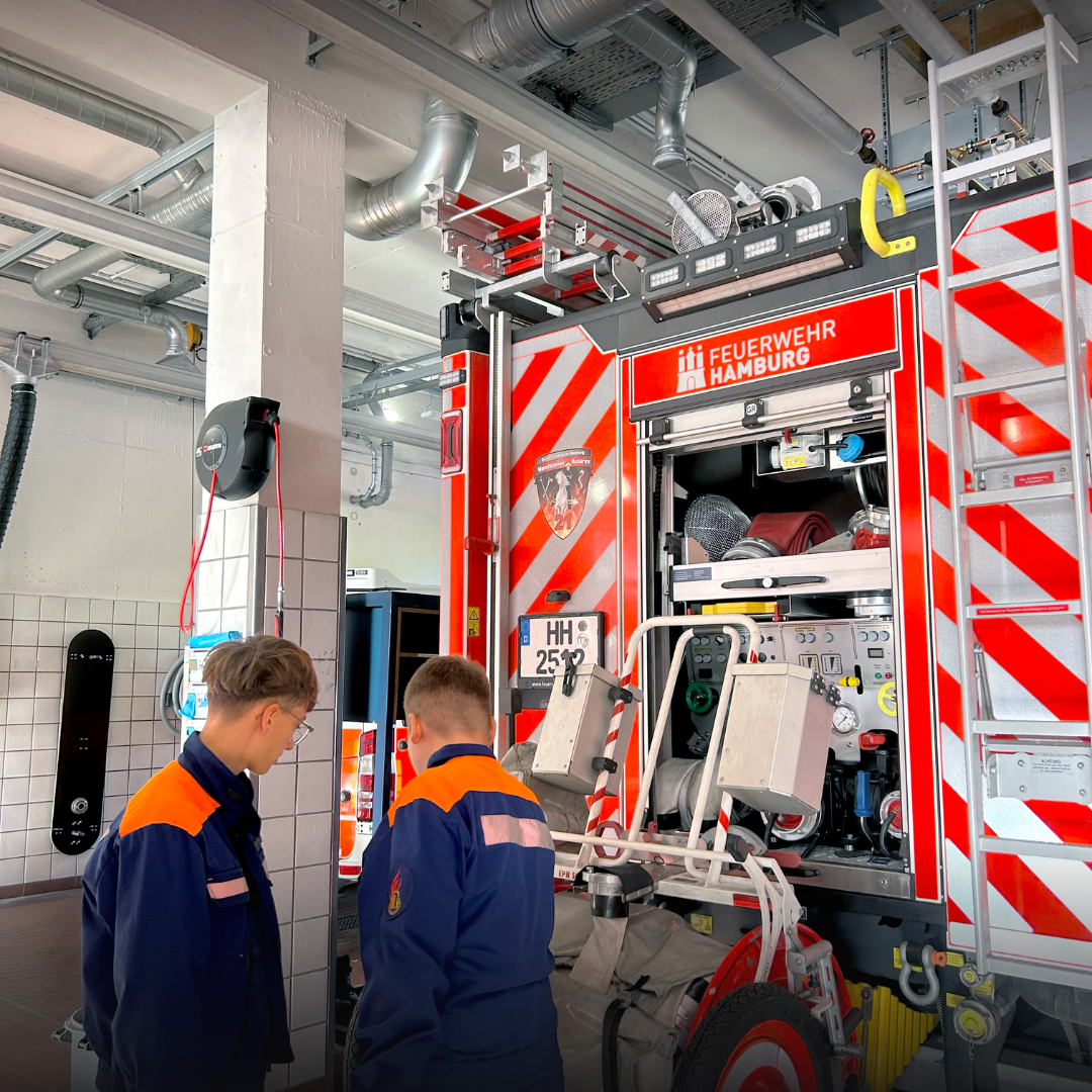 Zwei Jugendliche in Jugendfeuerwehruniform schauen sich das geöffnete Heck eines Löschfahrzeugs der Feuerwehr Hamburg mit Pumpe und Armaturen an.