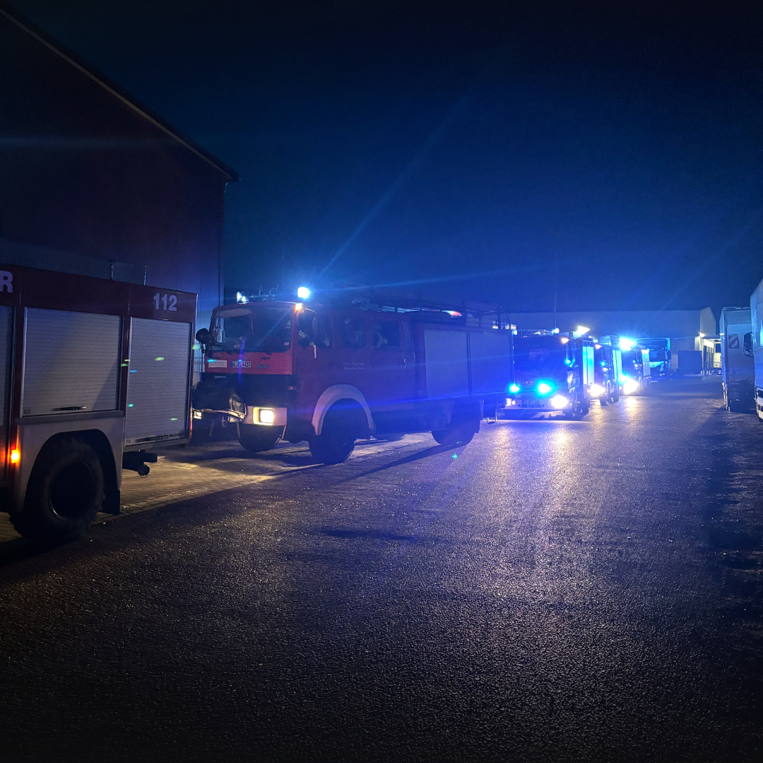 Mehrere Feuerwehrfahrzeuge mit eingeschaltetem Blaulicht stehen nachts hintereinander auf der Zufahrt neben einer Halle.