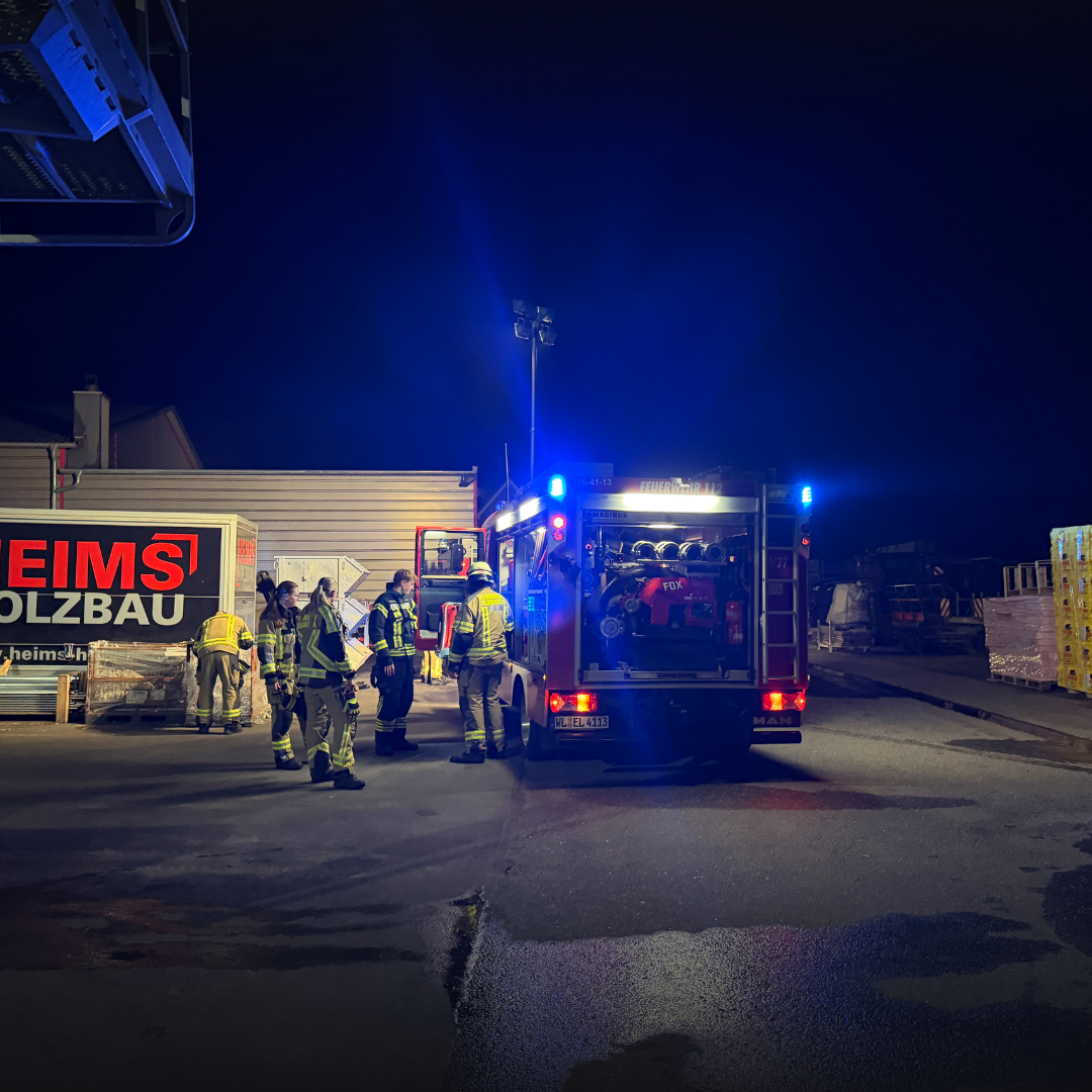 Löschfahrzeug mit Blaulicht steht nachts vor einer Lagerhalle, mehrere Einsatzkräfte arbeiten am Heck des Fahrzeugs.